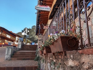 A picturesque street scene featuring a stone staircase lined with ornate iron railings and flower boxes filled with colorful blooms. Traditional buildings with wooden balconies and slanted roofs line the cobblestone street, adding to the quaint, charming atmosphere. Signs for a churro shop and other eateries are visible, suggesting a cozy, inviting environment.