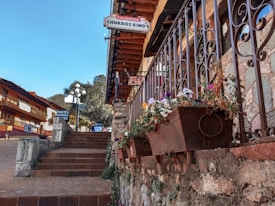 A picturesque street scene featuring a stone staircase lined with ornate iron railings and flower boxes filled with colorful blooms. Traditional buildings with wooden balconies and slanted roofs line the cobblestone street, adding to the quaint, charming atmosphere. Signs for a churro shop and other eateries are visible, suggesting a cozy, inviting environment.