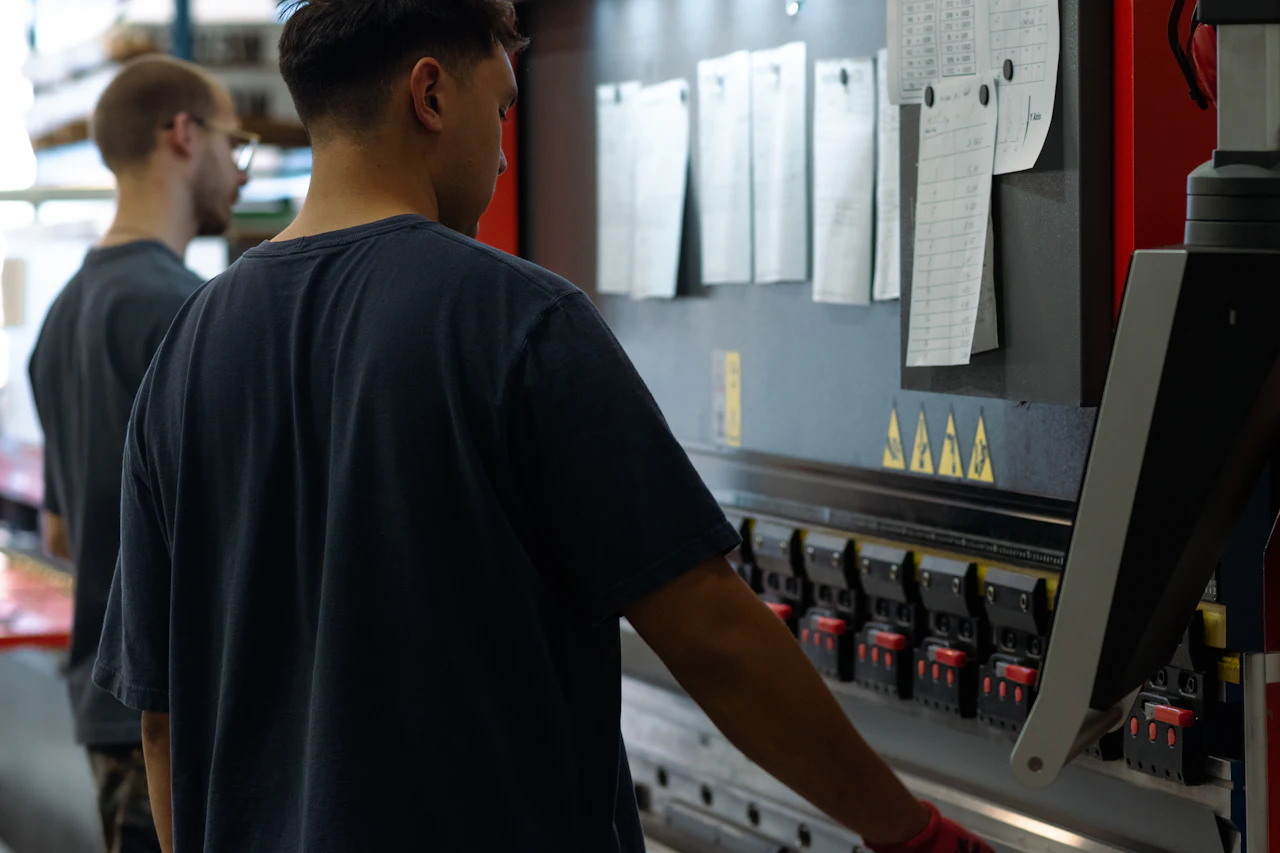 Two operators at a press brake; job sheets are pinned on the board above it.