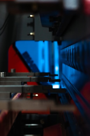 A close-up of industrial machinery with metallic components, featuring a combination of mechanical arms and a sleek, angular design. The background is infused with a bright blue light, contrasting with darker shadows and red accents.