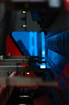 A close-up of industrial machinery with metallic components, featuring a combination of mechanical arms and a sleek, angular design. The background is infused with a bright blue light, contrasting with darker shadows and red accents.