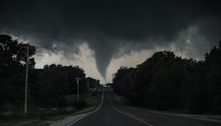 A large tornado spirals down from dark, swirling storm clouds over a rural road. The surrounding landscape is heavily wooded, with electrical poles lining the road. The sky is ominous, suggesting impending severe weather.
