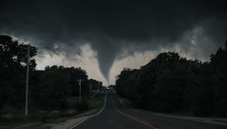 A large tornado spirals down from dark, swirling storm clouds over a rural road. The surrounding landscape is heavily wooded, with electrical poles lining the road. The sky is ominous, suggesting impending severe weather.