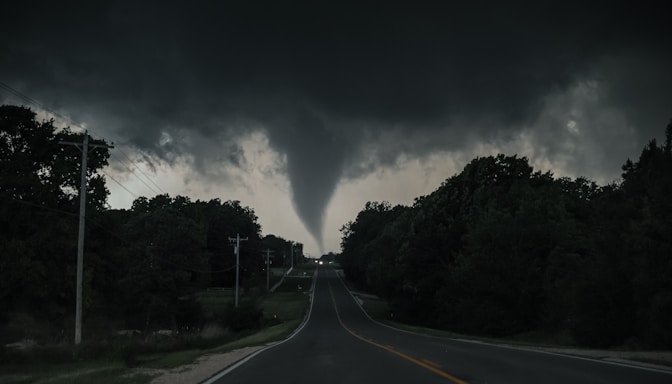 A large tornado spirals down from dark, swirling storm clouds over a rural road. The surrounding landscape is heavily wooded, with electrical poles lining the road. The sky is ominous, suggesting impending severe weather.