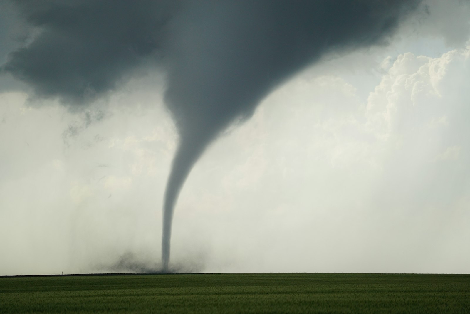 A dark funnel cloud touching down over a flat green field under a stormy sky