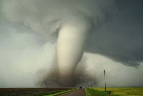 A large tornado funnel is touching down on a rural landscape, with dark, swirling storm clouds above. The tornado is surrounded by debris and dust near the ground. A road leads towards the tornado, with a vehicle traveling on it. Power lines run along the side of the road, disappearing into the distance.