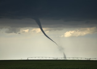 A tornado extends from dark, stormy clouds down to a flat, green field, with an irrigation system in the background. The sky is overcast with a foreboding atmosphere.