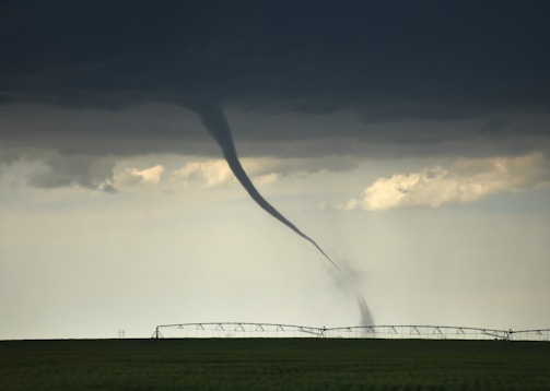 A tornado extends from dark, stormy clouds down to a flat, green field, with an irrigation system in the background. The sky is overcast with a foreboding atmosphere.