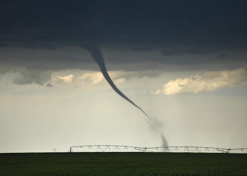 A tornado extends from dark, stormy clouds down to a flat, green field, with an irrigation system in the background. The sky is overcast with a foreboding atmosphere.
