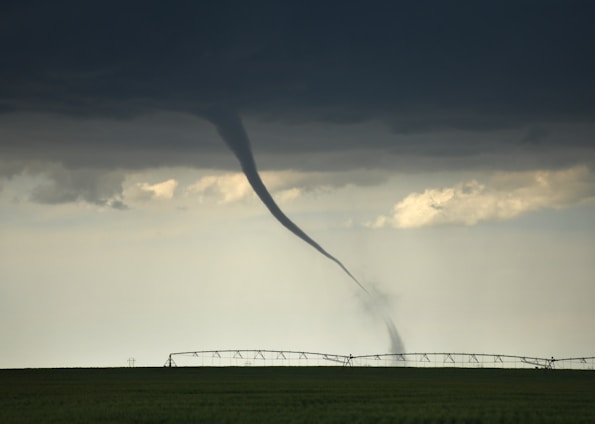 A tornado extends from dark, stormy clouds down to a flat, green field, with an irrigation system in the background. The sky is overcast with a foreboding atmosphere.