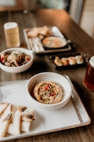 An assortment of dishes is arranged on a wooden table, featuring a prominent bowl of hummus garnished with red peppers and herbs, served with slices of grilled pita bread. In the background, there are bowls with various dishes, including a small black skillet with creamy soup and a platter of what appears to be dumplings.