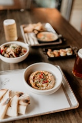 An assortment of dishes is arranged on a wooden table, featuring a prominent bowl of hummus garnished with red peppers and herbs, served with slices of grilled pita bread. In the background, there are bowls with various dishes, including a small black skillet with creamy soup and a platter of what appears to be dumplings.