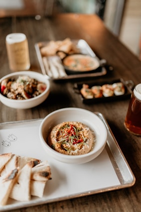 An assortment of dishes is arranged on a wooden table, featuring a prominent bowl of hummus garnished with red peppers and herbs, served with slices of grilled pita bread. In the background, there are bowls with various dishes, including a small black skillet with creamy soup and a platter of what appears to be dumplings.