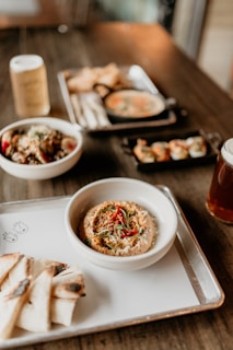 An assortment of dishes is arranged on a wooden table, featuring a prominent bowl of hummus garnished with red peppers and herbs, served with slices of grilled pita bread. In the background, there are bowls with various dishes, including a small black skillet with creamy soup and a platter of what appears to be dumplings.
