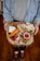 A charcuterie board held by a person, featuring various types of cheese, meats, a radish slice, green beans, carrots, and slices of grilled bread. The board has a rustic, natural wood finish, and the person is wearing a denim shirt.