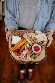 A charcuterie board held by a person, featuring various types of cheese, meats, a radish slice, green beans, carrots, and slices of grilled bread. The board has a rustic, natural wood finish, and the person is wearing a denim shirt.