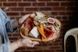A person holds a rustic wooden platter with an assortment of gourmet foods, including sliced cured meats, soft blue cheese, triangular bread pieces, carrot sticks, pickles, and a slice of watermelon radish. The background features a red brick wall, adding a cozy and warm atmosphere.