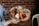 A person holds a rustic wooden platter with an assortment of gourmet foods, including sliced cured meats, soft blue cheese, triangular bread pieces, carrot sticks, pickles, and a slice of watermelon radish. The background features a red brick wall, adding a cozy and warm atmosphere.