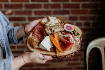A person holds a rustic wooden platter with an assortment of gourmet foods, including sliced cured meats, soft blue cheese, triangular bread pieces, carrot sticks, pickles, and a slice of watermelon radish. The background features a red brick wall, adding a cozy and warm atmosphere.