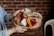 A person holds a rustic wooden platter with an assortment of gourmet foods, including sliced cured meats, soft blue cheese, triangular bread pieces, carrot sticks, pickles, and a slice of watermelon radish. The background features a red brick wall, adding a cozy and warm atmosphere.