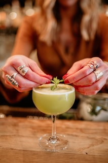 A person is delicately garnishing a frothy, light green cocktail with a small mint leaf. The cocktail is served in an elegant coupe glass resting on a wooden surface. The individual has several rings on their fingers and polished nails, adding a touch of style and sophistication.
