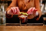 A person with red nail polish is garnishing a cocktail with a twist of lemon zest. The drink is in a short glass filled with ice and a dark liquid, placed on a bar counter. A bottle of ginger beer is visible to the left, and a measuring jigger is partially visible on the right.