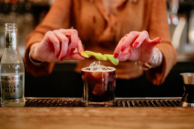 A person with red nail polish is garnishing a cocktail with a twist of lemon zest. The drink is in a short glass filled with ice and a dark liquid, placed on a bar counter. A bottle of ginger beer is visible to the left, and a measuring jigger is partially visible on the right.