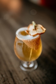 A glass of amber-colored beverage, possibly a cocktail or beer, is garnished with a dried apple slice. The drink is placed on a wooden surface, and soft lighting creates a warm, inviting atmosphere.