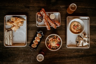 A variety of appetizers arranged on metal trays and wooden table, including wraps, chips, pretzels, hummus with toppings, and grilled flatbreads. Two glasses of beer accompany the dishes.