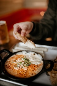 A person holds a piece of bread or cracker with a spread or dip on it, above a small cast iron skillet containing a baked dish topped with herbs and cheese. The dish appears creamy with a golden, crispy surface. The background is softly blurred, emphasizing the food in the foreground.