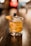A cocktail glass filled with a golden-brown beverage sits on a polished wooden surface. The drink is adorned with a clear ice cube, a twist of orange peel, and a dark cherry skewer. The background is a bokeh effect with warm, blurry lights, adding to an elegant and cozy atmosphere.