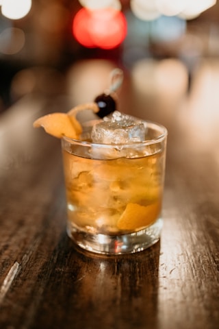 A cocktail glass filled with a golden-brown beverage sits on a polished wooden surface. The drink is adorned with a clear ice cube, a twist of orange peel, and a dark cherry skewer. The background is a bokeh effect with warm, blurry lights, adding to an elegant and cozy atmosphere.