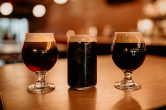 Three glasses of dark beer are placed on a wooden surface in a dimly lit bar or pub setting. Two of the glasses are goblets, and the third is a pint glass. The background is softly blurred, highlighting warm lights and a cozy atmosphere.