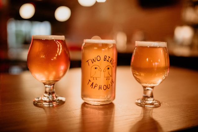 Three glasses of beer are arranged on a wooden surface. The central glass is a branded pint glass with the words 'Two Birds Tap House' alongside simple illustrations of two birds. The glass is flanked by two goblet-style beer glasses filled with beer, each showing a foamy head. The background has a warm, blurred ambiance with circular bokeh lights, suggesting a cozy bar or pub setting.