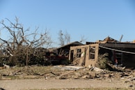 A scene of devastation with a partially collapsed building and a toppled tree. Debris is scattered across the area, and a damaged vehicle is partially visible. The sky is clear, contrasting the destruction on the ground.