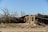 A scene of devastation with a partially collapsed building and a toppled tree. Debris is scattered across the area, and a damaged vehicle is partially visible. The sky is clear, contrasting the destruction on the ground.