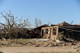 A scene of devastation with a partially collapsed building and a toppled tree. Debris is scattered across the area, and a damaged vehicle is partially visible. The sky is clear, contrasting the destruction on the ground.