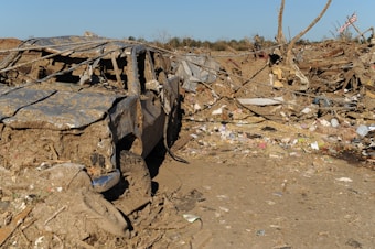 A scene depicting significant destruction, with a damaged vehicle covered in mud and debris in the foreground. The surrounding area is cluttered with broken pieces of wood, metal, and other wreckage. An American flag is visible amidst the chaos, suggesting a disaster or catastrophic event.