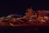 A large pile of debris consisting of wooden beams, metal, and household items scattered across the ground. The scene is dimly lit, indicating it could be nighttime or poorly illuminated. The debris appears to be the result of a destructive event, with materials tossed haphazardly, suggesting the remains of a building or home.