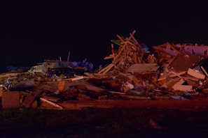 A large pile of debris consisting of wooden beams, metal, and household items scattered across the ground. The scene is dimly lit, indicating it could be nighttime or poorly illuminated. The debris appears to be the result of a destructive event, with materials tossed haphazardly, suggesting the remains of a building or home.