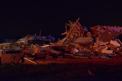 A large pile of debris consisting of wooden beams, metal, and household items scattered across the ground. The scene is dimly lit, indicating it could be nighttime or poorly illuminated. The debris appears to be the result of a destructive event, with materials tossed haphazardly, suggesting the remains of a building or home.