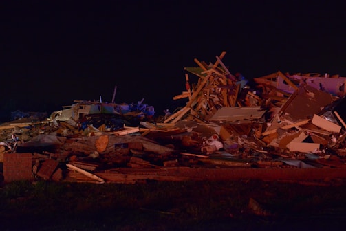 A large pile of debris consisting of wooden beams, metal, and household items scattered across the ground. The scene is dimly lit, indicating it could be nighttime or poorly illuminated. The debris appears to be the result of a destructive event, with materials tossed haphazardly, suggesting the remains of a building or home.