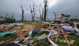 A scene of destruction and debris with splintered wood, scattered belongings, and damaged trees. The remnants of a structure, possibly a house, are partially visible amidst the wreckage. The sky is overcast, contributing to the somber atmosphere.
