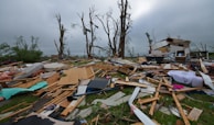 A scene of destruction and debris with splintered wood, scattered belongings, and damaged trees. The remnants of a structure, possibly a house, are partially visible amidst the wreckage. The sky is overcast, contributing to the somber atmosphere.