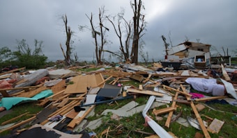 A scene of destruction and debris with splintered wood, scattered belongings, and damaged trees. The remnants of a structure, possibly a house, are partially visible amidst the wreckage. The sky is overcast, contributing to the somber atmosphere.