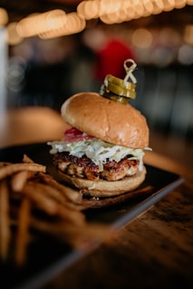 A delicious hamburger is placed on a dark plate, topped with slaw and two pickles skewered on a stick. Accompanying the burger are crispy fries, and the background is softly blurred with warm lighting and a cozy ambiance.