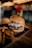 A delicious hamburger is placed on a dark plate, topped with slaw and two pickles skewered on a stick. Accompanying the burger are crispy fries, and the background is softly blurred with warm lighting and a cozy ambiance.