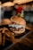A delicious hamburger is placed on a dark plate, topped with slaw and two pickles skewered on a stick. Accompanying the burger are crispy fries, and the background is softly blurred with warm lighting and a cozy ambiance.
