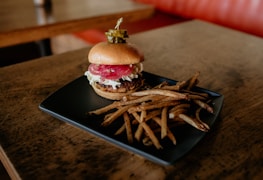 A hamburger with lettuce, onion, and cheese, topped with pickles on a toothpick, is placed next to a pile of fries on a black rectangular plate. The setting is a wooden table in a restaurant.