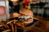 A gourmet burger with a juicy patty topped with lettuce, pickles, and onions sits on a dark plate. The burger is accompanied by a side of golden-brown French fries. The background is softly blurred, suggesting a cozy dining atmosphere.